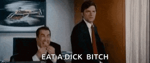 two men in suits and ties are standing next to each other in a room and one of them is eating a sandwich .