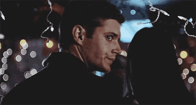a man in a suit is looking at a woman in a dark room with christmas lights in the background .