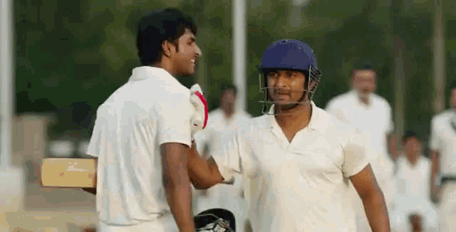 two cricket players are shaking hands on a field and one of them is wearing a helmet .