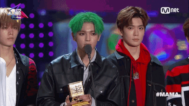 a man with green hair holds a trophy in front of a microphone at a mnet event