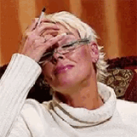 a woman wearing glasses and a white sweater is sitting on a couch covering her face with her hand .