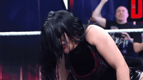 a woman in a black dress is kneeling down in a wrestling ring while a man watches .