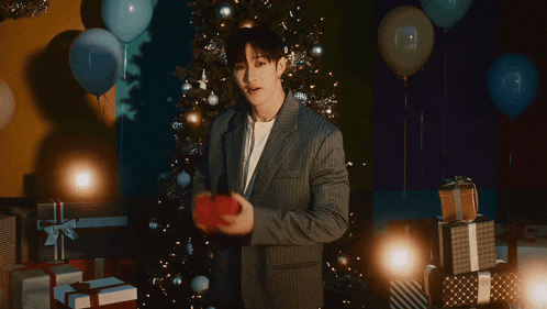 a man in a suit is holding a gift in front of a christmas tree and balloons