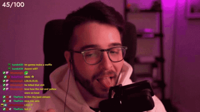 a man with glasses is licking his tongue while sitting in front of a microphone with the number 45/100 on the bottom right