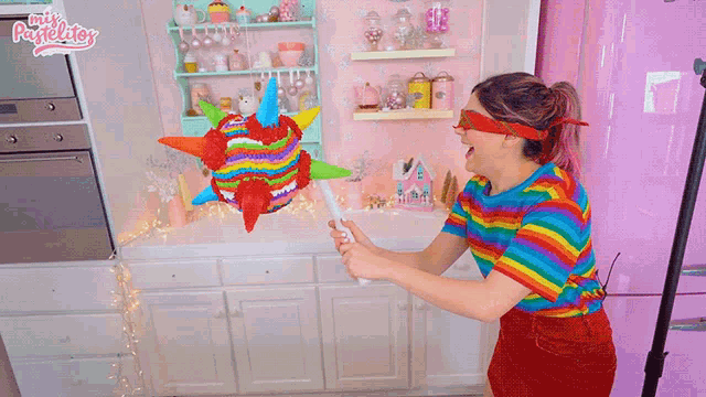 a woman in a rainbow shirt is holding a pinata in front of a pink refrigerator that says mis pastelitos