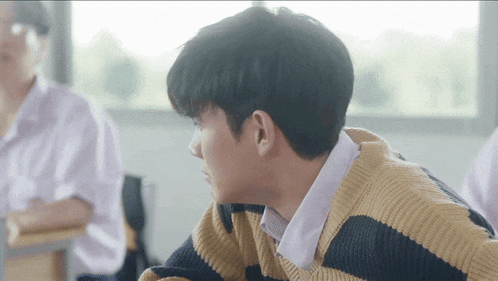a young man in a striped sweater is sitting at a desk in a classroom with other students .