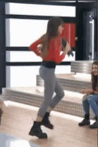 a woman in a red shirt and gray pants is dancing in a room with a microphone .