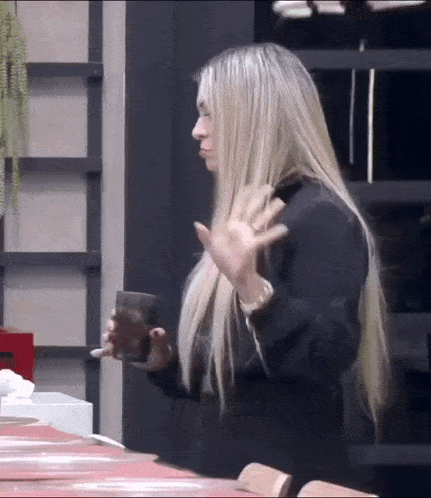 a woman with long blonde hair is standing at a table holding a cup of coffee .