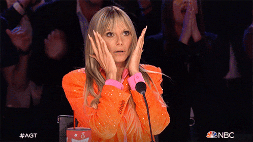 a woman in an orange jacket stands in front of a microphone with her hands on her face and a nbc logo behind her