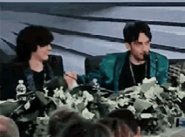 two men are sitting at a table talking into microphones while a crowd watches .