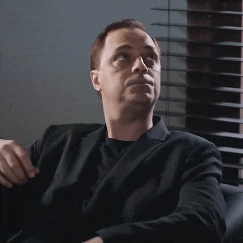 a man in a suit sits in front of a window with blinds