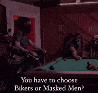a man riding a motorcycle next to a pool table with a masked man playing pool .
