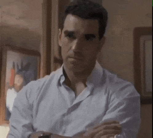a man in a white shirt is sitting in a room with his arms crossed and looking at the camera .