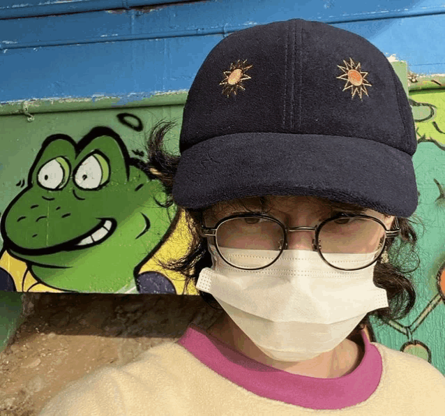 a person wearing a face mask and glasses stands in front of a wall with a frog on it