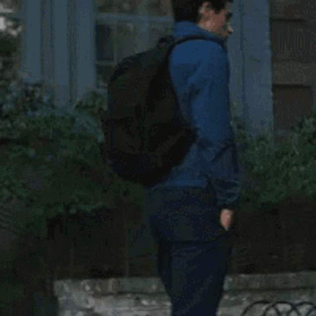 a man wearing a blue jacket and a backpack stands in front of a blue building .