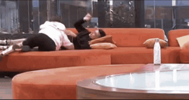two people are laying on an orange couch in a living room with a glass table in the foreground