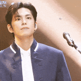 a man in a blue jacket stands in front of a microphone with the word be visible in the background