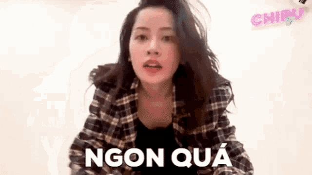 a woman in a plaid shirt is standing in front of a white wall and talking .