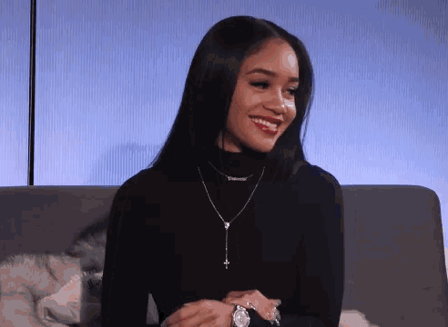 a woman wearing a black turtleneck and a rosary necklace is smiling while sitting on a couch