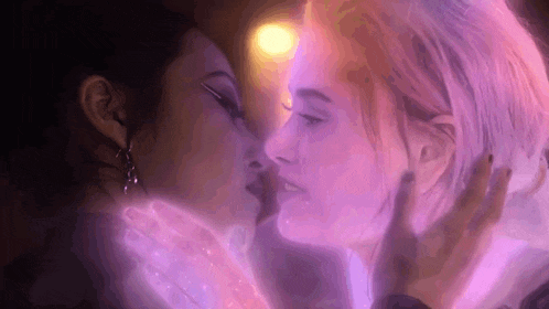 two women are kissing each other in a dark room with purple lights behind them .