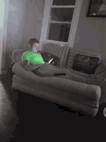 a man in a green shirt sits on a couch looking at his phone