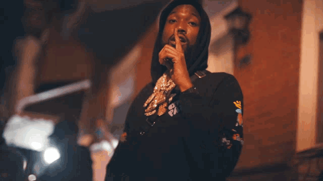 a man wearing a black hoodie with a crown on the sleeve holds his finger to his lips