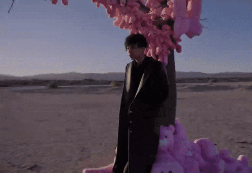 a man in a black coat is standing in front of a tree covered in pink stuffed animals