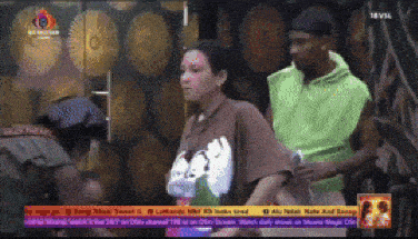 a man in a green hoodie stands next to a woman in a brown shirt on a television screen