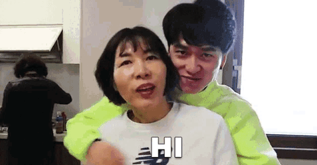 a man and a woman are hugging each other in a kitchen . the man is wearing a neon green sweater .