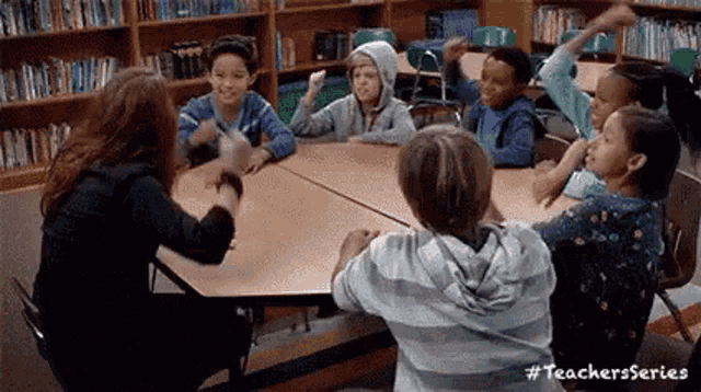 a group of children are sitting around a table in a library with their arms in the air .