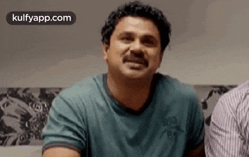 a man with a mustache is wearing a green shirt and smiling while sitting on a couch .