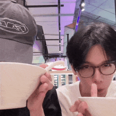 a man wearing glasses is eating from a bowl while another man wearing a hat with the letter e on it watches