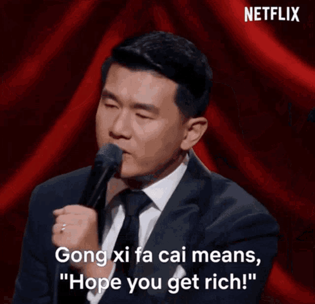 a man in a suit and tie is speaking into a microphone and saying " gong xi fa cai means hope you get rich "