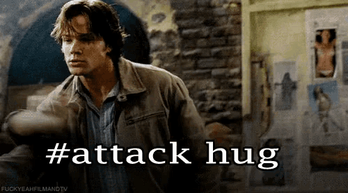 a man in a brown jacket stands in front of a brick wall with the words #attack hug below him