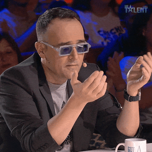 a man wearing sunglasses and a suit sits at a table in front of a crowd and the word talent is on the bottom