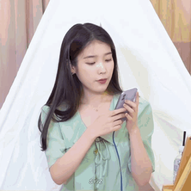 a woman in a green top is holding a cell phone with syiz written on the bottom right