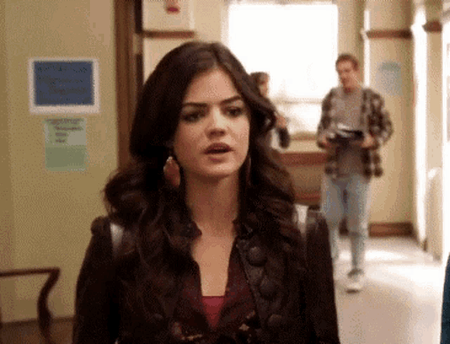 a woman in a leather jacket is walking down a hallway with a man in a plaid shirt in the background .