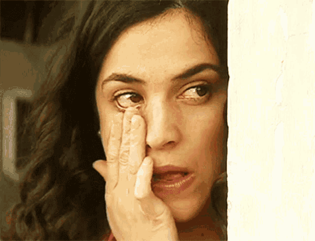 a woman wipes her eye with her hand while looking out a window