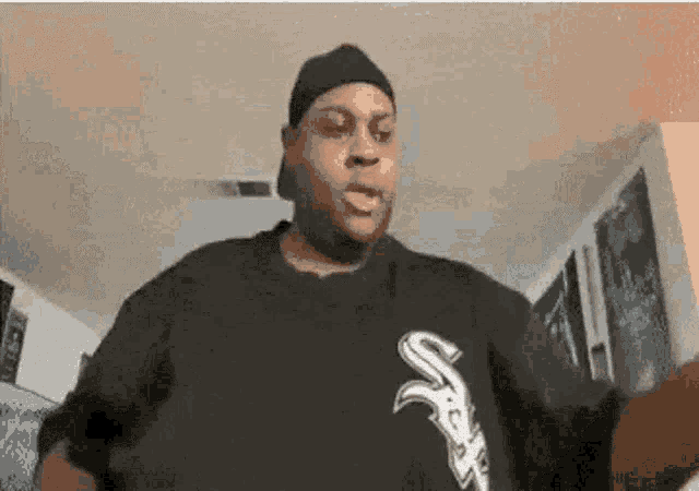 a man wearing a hat and a black shirt with the letter s on it is standing in a living room .