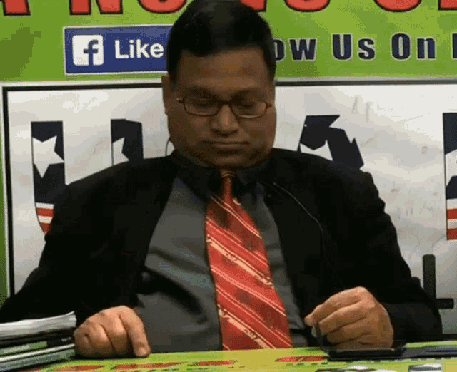 a man in a suit and tie sits in front of a sign that says like