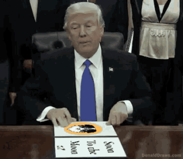 donald trump is sitting at a table with a sign on it that says " send son to the moon "
