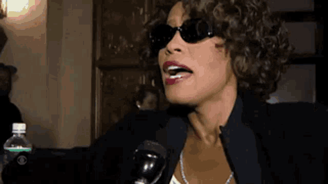 a woman wearing sunglasses and a black jacket is speaking into a microphone .