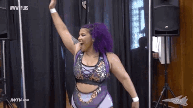 a woman with purple hair is standing in front of a black curtain with her arm in the air .