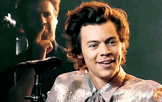 harry styles is smiling in front of a microphone while another man looks on .