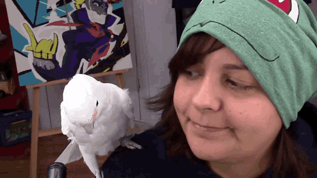 a woman wearing a green beanie looks at a white parrot on her shoulder