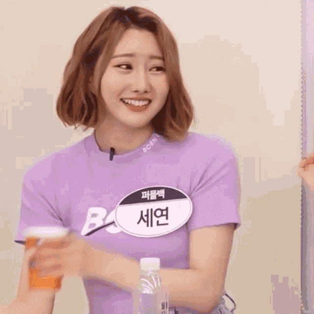 a woman in a purple shirt is smiling and holding a cup and a bottle .