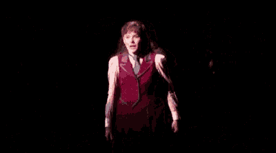 a woman is standing on a stage in a dark room wearing a red vest and tie .