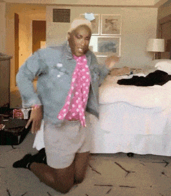 a man in a denim jacket and pink scarf is kneeling on the floor in a bedroom