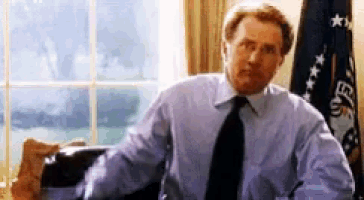 a man in a blue shirt and tie sits at a desk in front of a flag that says ed