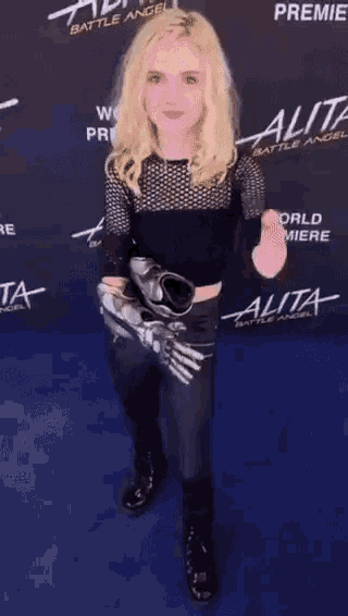 a woman with blonde hair is standing on a blue carpet in front of a sign that says alita